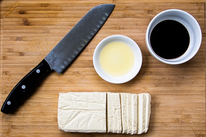 Tofu block on cutting board with knife, sesame oil and soy sauce